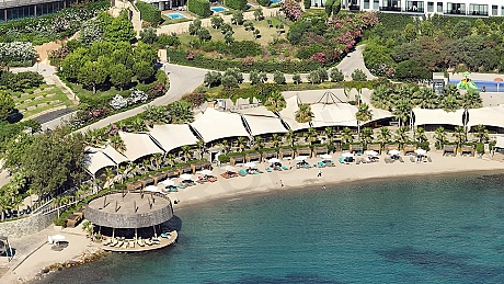 Bodrum'da özel bir rezidans plajı, berrak turkuaz denizi ve ince kumları ile güneşli bir manzara. Plajda şezlonglar ve şemsiyeler düzenli bir şekilde yer alıyor.