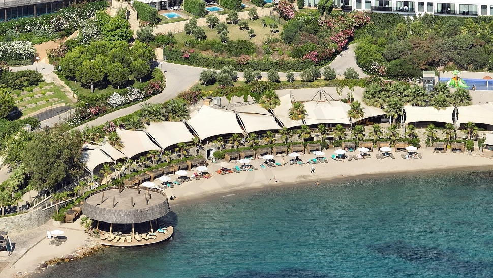 Bodrum'da özel bir rezidans plajı, berrak turkuaz denizi ve ince kumları ile güneşli bir manzara. Plajda şezlonglar ve şemsiyeler düzenli bir şekilde yer alıyor.