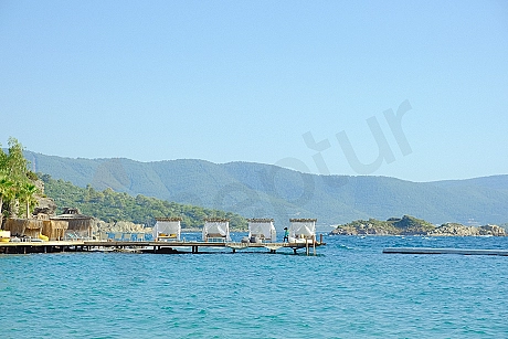 Deniz üzerinde uzanan ahşap bir özel iskele. İskele, güneşli bir havada sakin denize doğru uzanmakta ve çevresinde açık mavi deniz suyu görünmektedir.