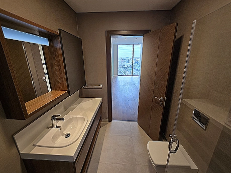 Modern tasarımlı, aydınlık ve geniş banyo iç mekanı. Beyaz seramikler, şık armatürler ve büyük bir ayna detayı.