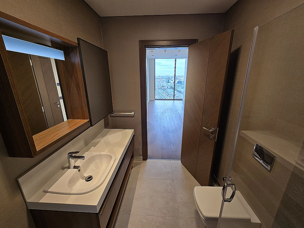 Modern tasarımlı, aydınlık ve geniş banyo iç mekanı. Beyaz seramikler, şık armatürler ve büyük bir ayna detayı.